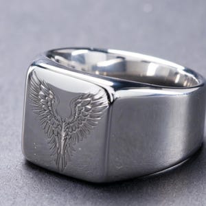 May include: A polished silver-tone signet ring featuring a flat, square-shaped face. The center of the face is engraved with a detailed design of symmetrical, feathered wings extending upwards and outwards, creating a distinct winged motif.