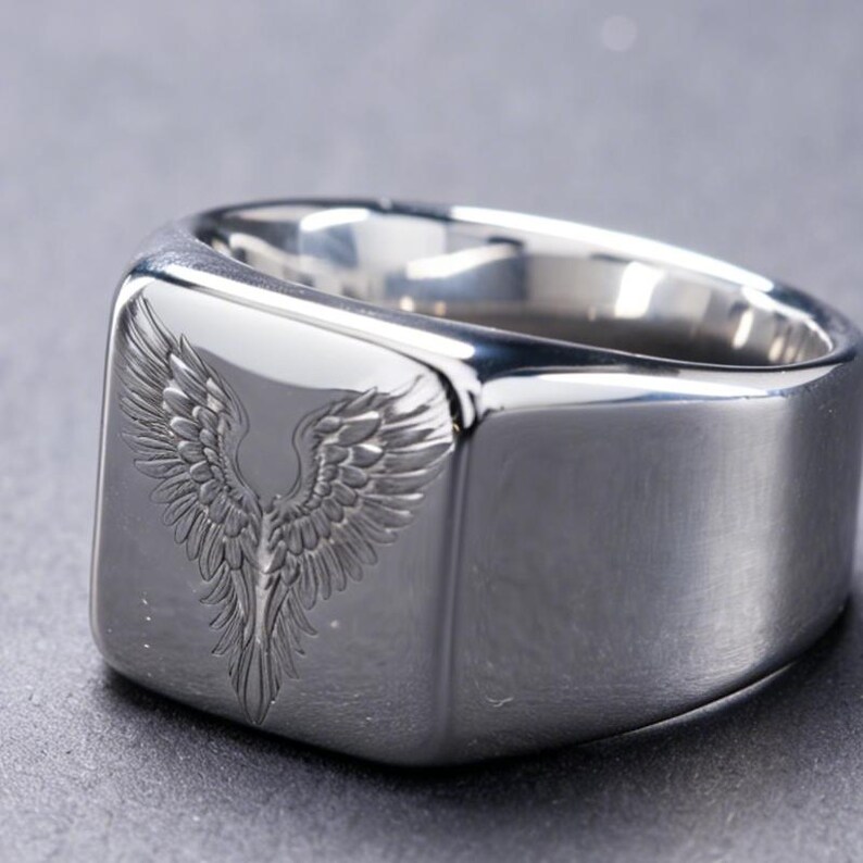 May include: A polished silver-tone signet ring featuring a flat, square-shaped face. The center of the face is engraved with a detailed design of symmetrical, feathered wings extending upwards and outwards, creating a distinct winged motif.
