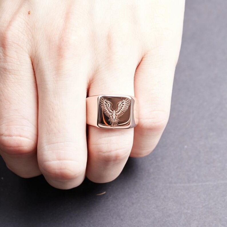 May include: A rose gold-toned signet ring featuring a square face with an engraved design of a bird with outstretched wings. The metal surface is polished and reflective, showcasing the detailed lines of the avian motif centered on the band.