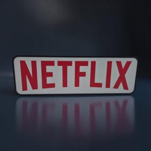 Può includere: Un cartello rettangolare con la scritta "NETFLIX" in lettere rosse in grassetto su sfondo bianco. Il cartello ha un bordo nero ed è posto su uno sfondo blu scuro. L'immagine è ben illuminata, evidenziando il design del cartello.