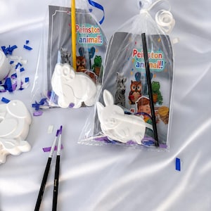 Pode incluir: Duas sacolas de celofane transparente amarradas com fitas azuis, cada uma contendo um artesanato branco em forma de animal e um cartão com o texto "Peinston animal!!". Os cartões apresentam animais de desenho animado. Pincéis e confetes também são visíveis.