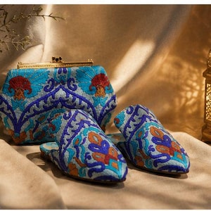 May include: A clutch and matching slippers with intricate embroidery in shades of blue, orange, and red. The clutch has a gold clasp. A decorative lantern is in the background, casting a warm glow.