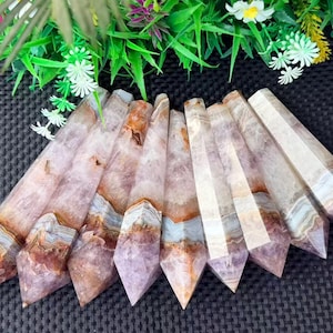 May include: A collection of seven polished crystal points arranged in a fan shape. Each stone features a hexagonal prism structure with a pointed tip, displaying a blend of translucent purple, white, and brown banded layers throughout the surface.