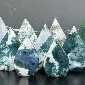 May include: A collection of nine teardrop-shaped polished stone carvings featuring various patterns of moss agate and banded agate. The stones display shades of deep forest green, white, translucent gray, and earthy brown with unique mineral inclusions.