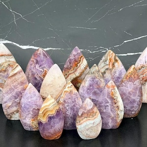 May include: A collection of teardrop-shaped polished stones featuring a mix of purple amethyst and banded agate patterns. The stones display natural layers of white, orange, and brown mineral deposits, with crystalline textures visible throughout.