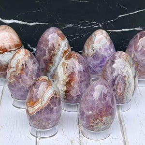 May include: A collection of eight polished, egg-shaped stones featuring banded patterns in shades of purple, white, and brown. Each stone is displayed on a small, clear circular stand, showcasing crystalline textures and smooth, glossy surfaces.