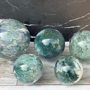 May include: Five polished moss agate spheres of varying sizes, each displaying unique green and white dendritic inclusions and patterns. The stones are smooth, rounded, and exhibit a glossy finish, showcasing natural mineral formations in shades of forest green, sage, and translucent white throughout their surfaces.