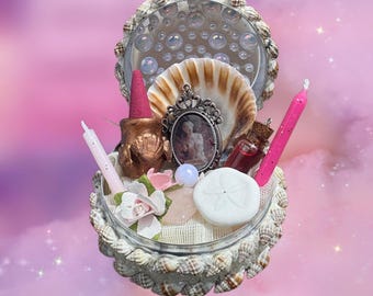 Puede incluir: Un recipiente redondo decorado con pequeñas conchas marinas que contiene un cono de incienso rosa, un caldero de color bronce, una vela rosa, un dólar de arena blanco, un pequeño frasco de vidrio con líquido rojo, un colgante y flores artificiales.