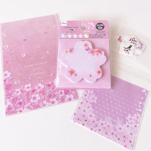 Op de afbeelding: Een verzameling roze briefpapier met kersenbloesemmotieven. Bevat een notitieblok met de tekst "CHERRY BLOSSOMS are in full bloom", een bloemvormig plaknotitieblok en papier met patronen.