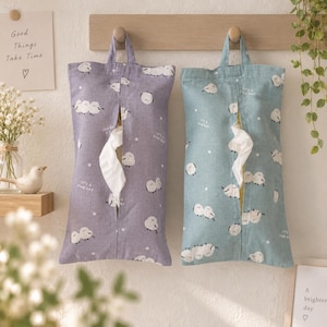 May include: Two rectangular fabric tissue holders, one purple and one light blue, each featuring a repeating pattern of small white sheep and the text "It's a fine day." Both holders have a central vertical slit for dispensing tissues.