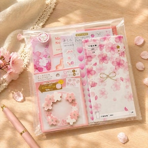 Puede incluir: Un conjunto de artículos de papelería con temática de flores de cerezo, empaquetado en una bolsa de plástico transparente. El conjunto incluye sobres rosas, pegatinas y papel de carta. Se ve un bolígrafo rosa con detalles dorados.