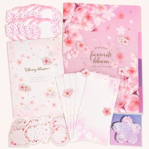 以下が含まれることがあります： ピンクと白の桜のモチーフが描かれたステーショナリーセット。「Find your favorite bloom in the warm of spring」と書かれたフォルダー、「Cherry blossom」と書かれたメモ帳、封筒、そして様々な花の形をした付箋が含まれています。