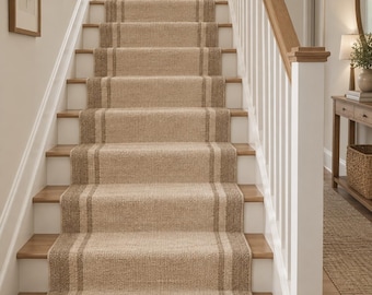 May include: A woven stair runner featuring a light tan centre panel bordered by darker brown stripes. The runner covers the centre of a wooden staircase with white risers and a wooden handrail, extending from the bottom step to the top landing.