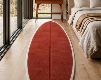 May include: An elongated, oval-shaped area rug featuring a deep terracotta red centre with a prominent vertical seam running down the middle, bordered by a thick, textured cream-coloured trim that follows the entire perimeter of the rug.