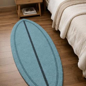 May include: An oval-shaped rug in a light blue colour with a dark blue border and a central dark blue vertical stripe. The rug features a pointed top and a notched bottom edge, mimicking the shape of a surfboard, with a textured pile surface.