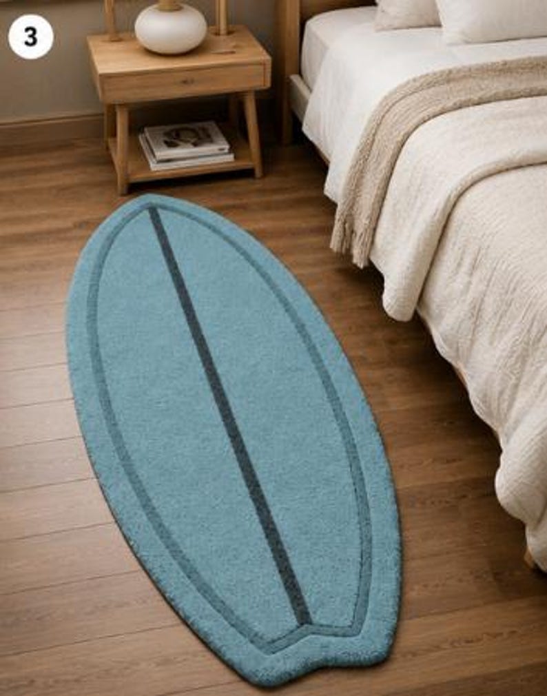 May include: An oval-shaped rug in a light blue colour with a dark blue border and a central dark blue vertical stripe. The rug features a pointed top and a notched bottom edge, mimicking the shape of a surfboard, with a textured pile surface.