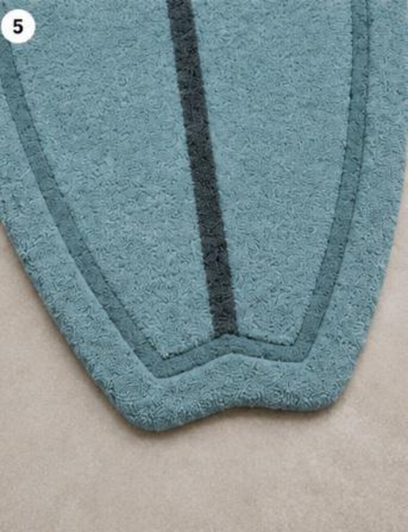 May include: A surfboard-shaped rug with a textured, light blue pile. The rug features a dark charcoal grey vertical stripe running down the centre and a matching dark charcoal grey border that follows the curved outline of the shape. The number 5 is visible in the top left corner.