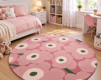 May include: A round area rug featuring a repeating pattern of large, stylised pink flowers with white centres and dark green stems. The rug has a textured, plush surface and is positioned on a light-coloured wooden floor in a bedroom setting.