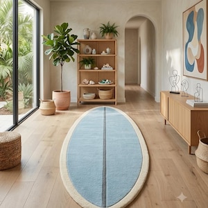 May include: An oval-shaped area rug featuring a light blue center with a dark gray vertical stripe down the middle, surrounded by a thick cream-colored border. The rug is positioned on a light-toned wooden floor in a bright, open interior space.