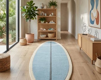 May include: An oval-shaped area rug featuring a light blue centre with a dark grey vertical stripe down the middle, surrounded by a thick cream-coloured border. The rug is positioned on a light-toned wooden floor in a bright, open interior space.