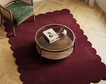 May include: A rectangular area rug in a deep burgundy colour featuring a scalloped edge design. The rug is made of a solid-coloured textile material and is positioned on a light-coloured wooden floor with a herringbone pattern.