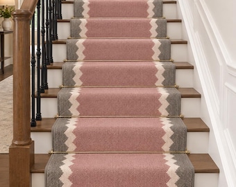 May include: A staircase runner featuring a woven texture with a central dusty pink panel flanked by grey borders and a white zigzag pattern. The runner is secured to wooden steps with metallic gold rods, contrasting with a black metal handrail.