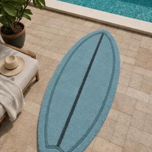 May include: A light blue rug shaped like a surfboard with a dark blue centre stripe and a matching dark blue border outline. The rug has a textured surface and is positioned vertically on a light-coloured stone patio surface.