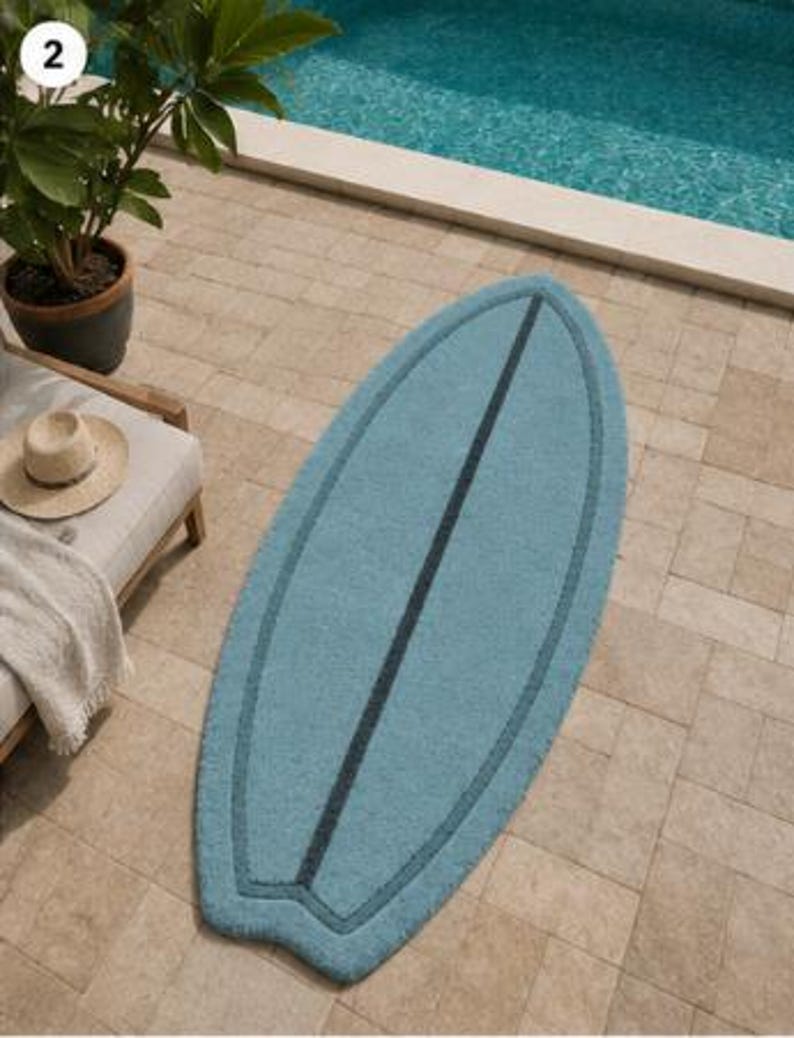 May include: A light blue rug shaped like a surfboard with a dark blue centre stripe and a matching dark blue border outline. The rug has a textured surface and is positioned vertically on a light-coloured stone patio surface.