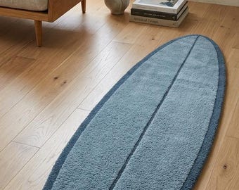 May include: An elongated, surfboard-shaped area rug featuring a textured, tufted pile surface. The rug is primarily light blue with a darker blue border and a thin, dark blue vertical line running down the centre, mimicking a classic surfboard design.
