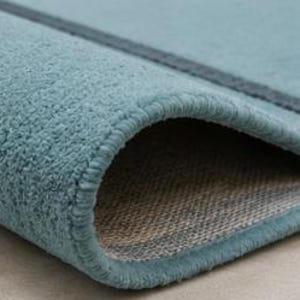 May include: A teal-coloured rug with a textured, plush surface and a dark teal stripe detail. The rug is folded over to reveal a woven, light-coloured backing and a finished, stitched edge. The number 7 is visible in the top left corner.