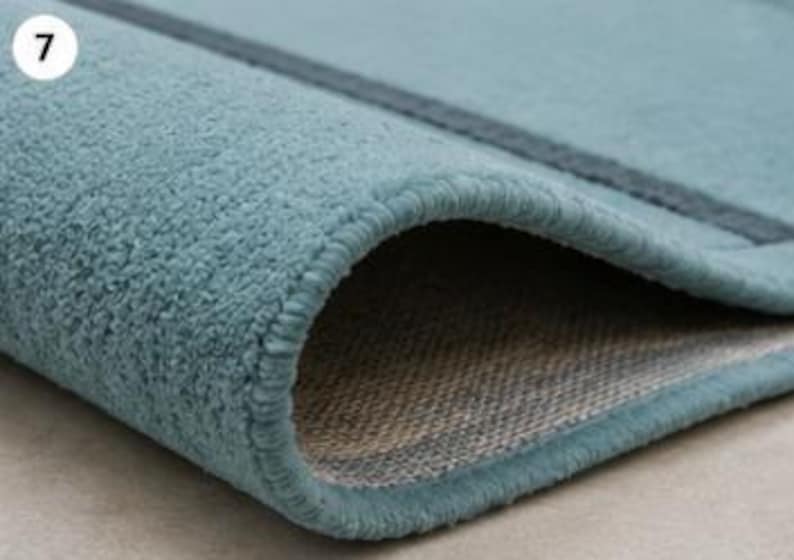 May include: A teal-coloured rug with a textured, plush surface and a dark teal stripe detail. The rug is folded over to reveal a woven, light-coloured backing and a finished, stitched edge. The number 7 is visible in the top left corner.