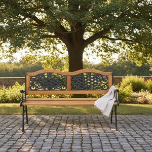May include: A wooden park bench with decorative black metal accents and legs. The bench has a light-colored wood seat and back with ornate metalwork. A folded white blanket rests on the bench. The bench is set on a brick patio under a large tree.
