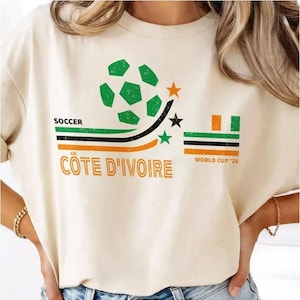 May include: A cream-colored t-shirt featuring a graphic with a green soccer ball, three stars, and horizontal stripes in green, black, and orange. The text reads SOCCER CÔTE D'IVOIRE and WORLD CUP '26. The design has a distressed, vintage-style texture.