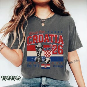 Puede incluir: Una camiseta de manga corta gris carb&oacute;n con un estampado gr&aacute;fico que incluye el texto World Cup CROATIA '26. El dise&ntilde;o presenta una ilustraci&oacute;n de un trofeo, un bal&oacute;n de f&uacute;tbol, un escudo a cuadros y una peque&ntilde;a bandera croata con el texto Est. 1912 Heritage.