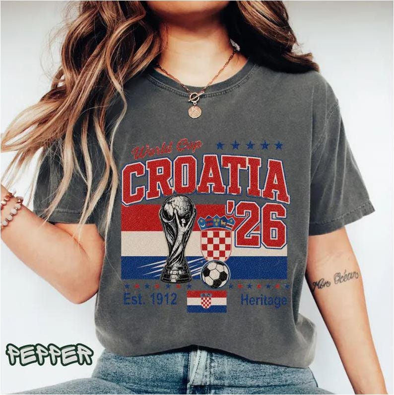 Puede incluir: Una camiseta de manga corta gris carb&oacute;n con un estampado gr&aacute;fico que incluye el texto World Cup CROATIA '26. El dise&ntilde;o presenta una ilustraci&oacute;n de un trofeo, un bal&oacute;n de f&uacute;tbol, un escudo a cuadros y una peque&ntilde;a bandera croata con el texto Est. 1912 Heritage.