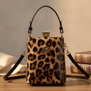 Puede incluir: Un bolso de mano tipo caja estructurado con un patrón de estampado de leopardo brillante en tonos marrón, beige y negro. El bolso incluye un asa superior, una correa de hombro desmontable, un cierre dorado y un emblema circular dorado con la letra M.