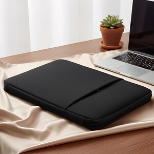 May include: A black laptop sleeve with a zippered pocket, resting on a beige satin surface. A laptop and a small potted succulent are in the background. The sleeve is designed to protect a laptop.