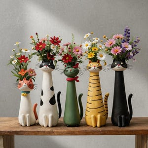 Puede incluir: Cinco jarrones con forma de gato en varios colores, cada uno con un ramo de flores. Los jarrones son blancos, negros, verdes, amarillos y una mezcla de naranja y blanco. Las flores incluyen margaritas, flores rojas y flores moradas. Los jarrones están sobre una superficie de madera.