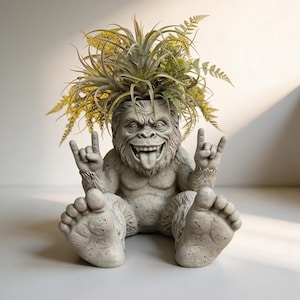 May include: A grey, concrete-effect planter shaped like a grinning, hairy creature with its tongue out and hands making a rock-and-roll gesture. The planter is filled with green and yellow plants, creating a unique decorative piece.