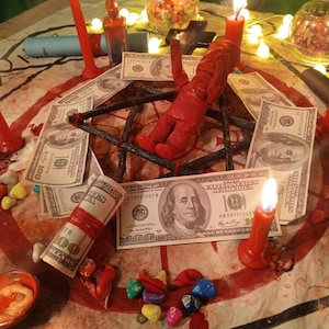 May include: A red doll placed in the center of a black star-shaped frame surrounded by several one-hundred-dollar bills, lit red candles, small glass bowls containing colorful stones, and a small glass bottle on a patterned fabric surface.