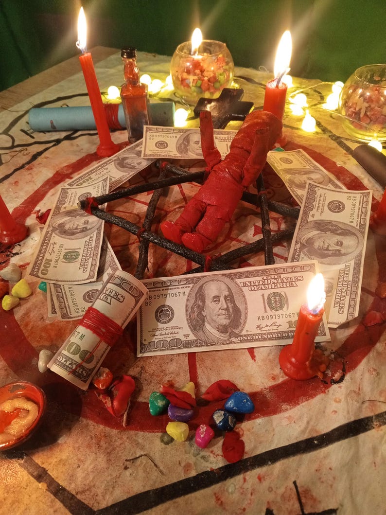 May include: A red doll placed in the center of a black star-shaped frame surrounded by several one-hundred-dollar bills, lit red candles, small glass bowls containing colorful stones, and a small glass bottle on a patterned fabric surface.