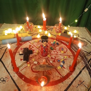 May include: A ritual setup featuring a central wooden doll, a black cross, and a leaf inscribed with the text "Your Name" on a circular cloth with red symbols. The arrangement is surrounded by red candles, small colorful stones, and fairy lights.