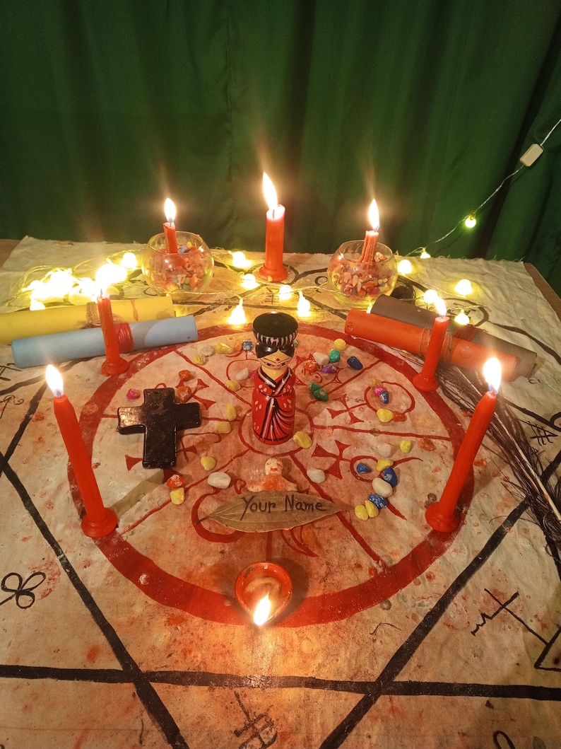 May include: A ritual setup featuring a central wooden doll, a black cross, and a leaf inscribed with the text "Your Name" on a circular cloth with red symbols. The arrangement is surrounded by red candles, small colorful stones, and fairy lights.