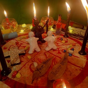 May include: A ritual setup featuring two white clay figures, a black figurine holding a scythe, and several lit candles. A circular cloth with red markings displays two leaves inscribed with the text Your Name and Partner Name.