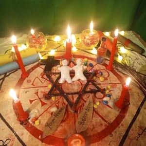 May include: A ritual setup featuring two white clay figures placed in the center of a red star drawn on fabric. Six lit red candles surround the figures, alongside two glass bowls, a small bottle, a black cross, and two leaves labeled Your Name.
