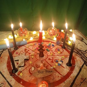 May include: A circular cloth featuring a red geometric design and the text Your Name on a leaf, surrounded by six black candles, one red candle, a small brown figurine, a cross, colorful stones, two glass bowls, and a small glass bottle.