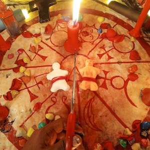 May include: A circular cloth featuring red occult symbols and a central burning red candle. Two small clay figures, one white and one orange, are tied with red string. A pair of red scissors is positioned between the figures on the cloth.