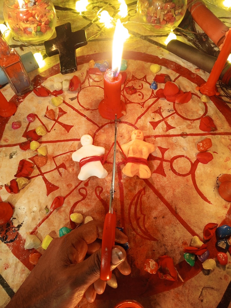 May include: A circular cloth featuring red occult symbols and a central burning red candle. Two small clay figures, one white and one orange, are tied with red string. A pair of red scissors is positioned between the figures on the cloth.