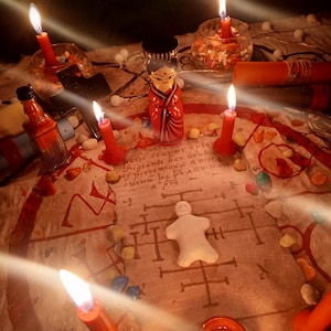 May include: A ritualistic arrangement featuring a central white clay figurine on a parchment with handwritten text and symbols, surrounded by red candles, a small wooden doll, a black cross, glass containers, and scattered colorful stones.