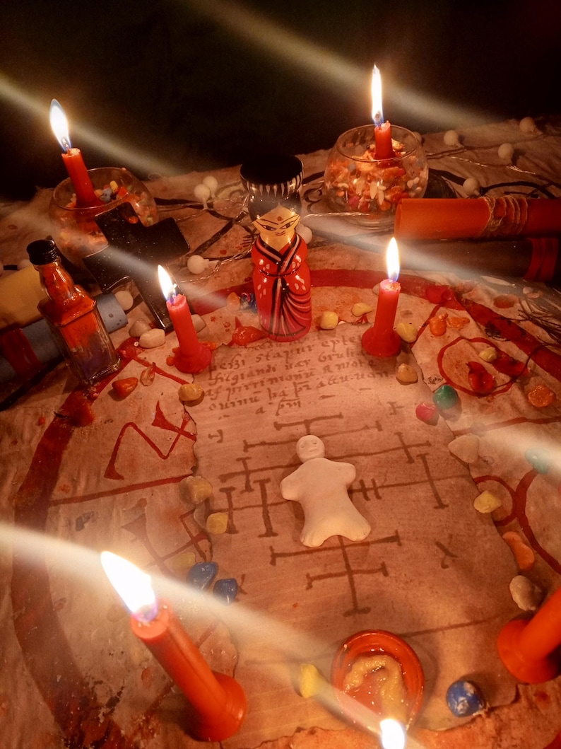 May include: A ritualistic arrangement featuring a central white clay figurine on a parchment with handwritten text and symbols, surrounded by red candles, a small wooden doll, a black cross, glass containers, and scattered colorful stones.